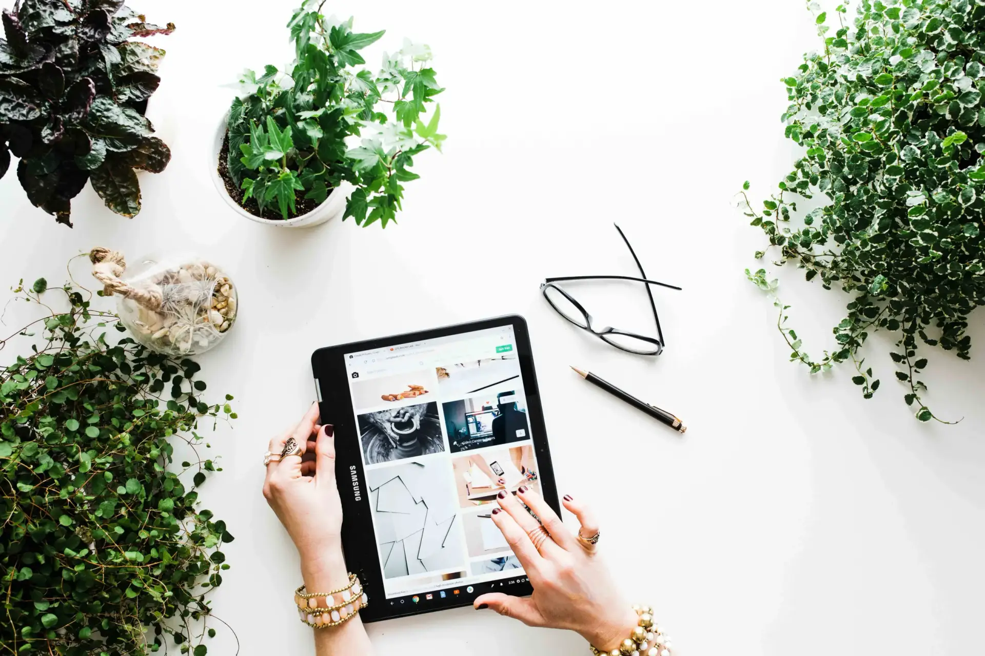 Woman's hands holding an iPad with multiple images opened on the screen. White desk with plants on the background.