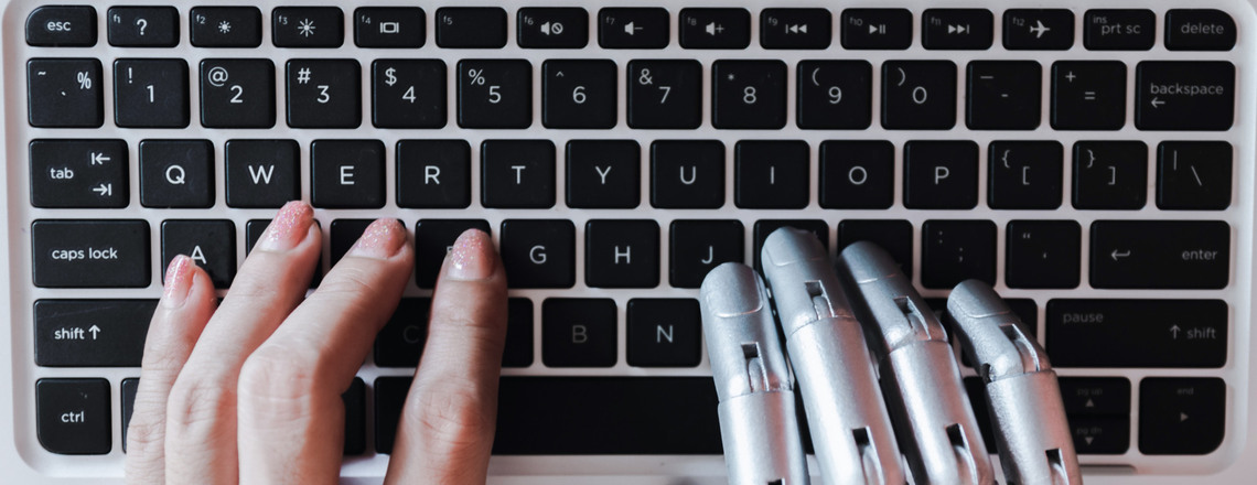 A human hand and a robotic hand typing together on a keyboard, symbolizing collaboration in creating E-E-A-T SEO content.