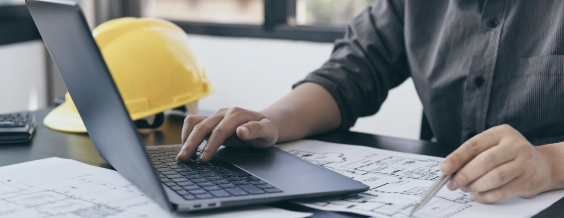 A person reviews blueprints while using a laptop at a desk with a yellow hard hat nearby, illustrating AI in the trades.